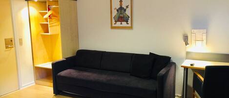A room with a dark-colored sofa, a small desk with a chair, a wooden cabinet, and a framed picture on the wall.