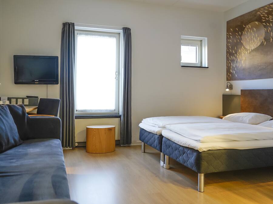 Una habitación de hotel con cama, sofá, televisión y ventana con cortinas.