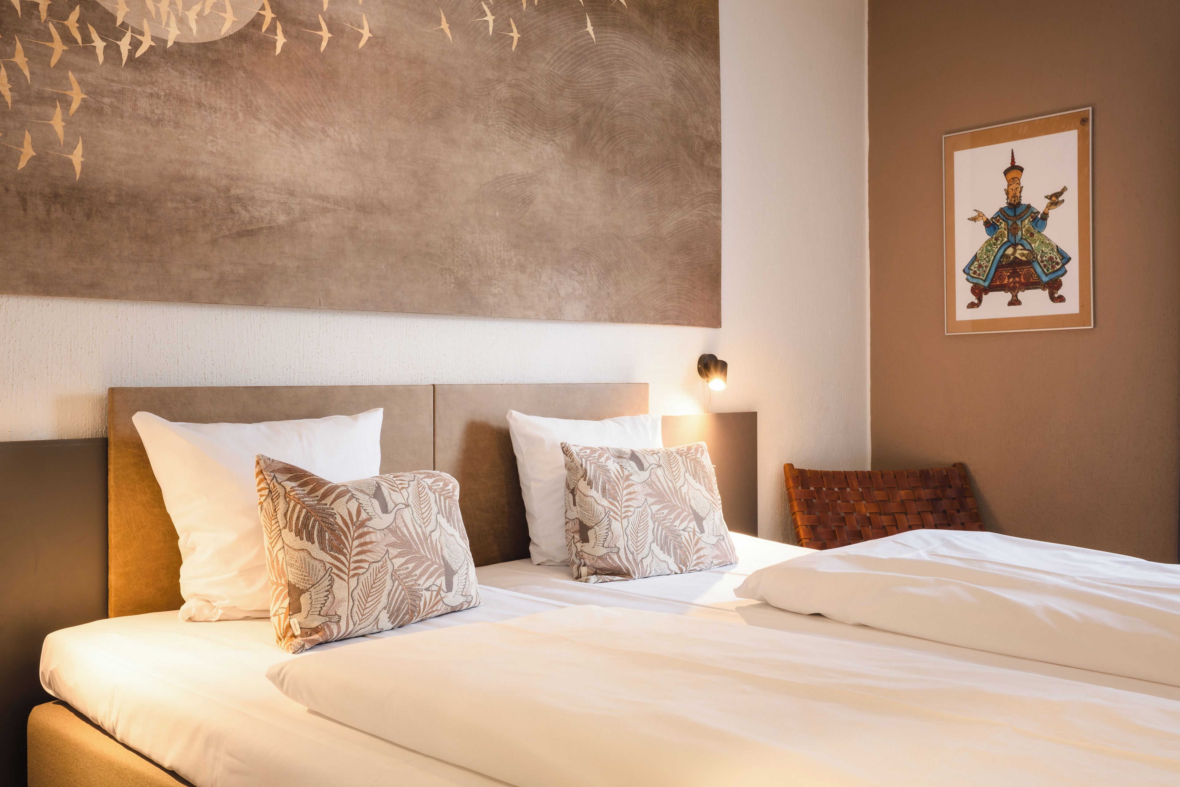 Une chambre d’hôtel avec un grand lit, deux oreillers à motifs de feuilles, une tête de lit, une lampe fixée au mur et un tableau encadré accroché au mur.
