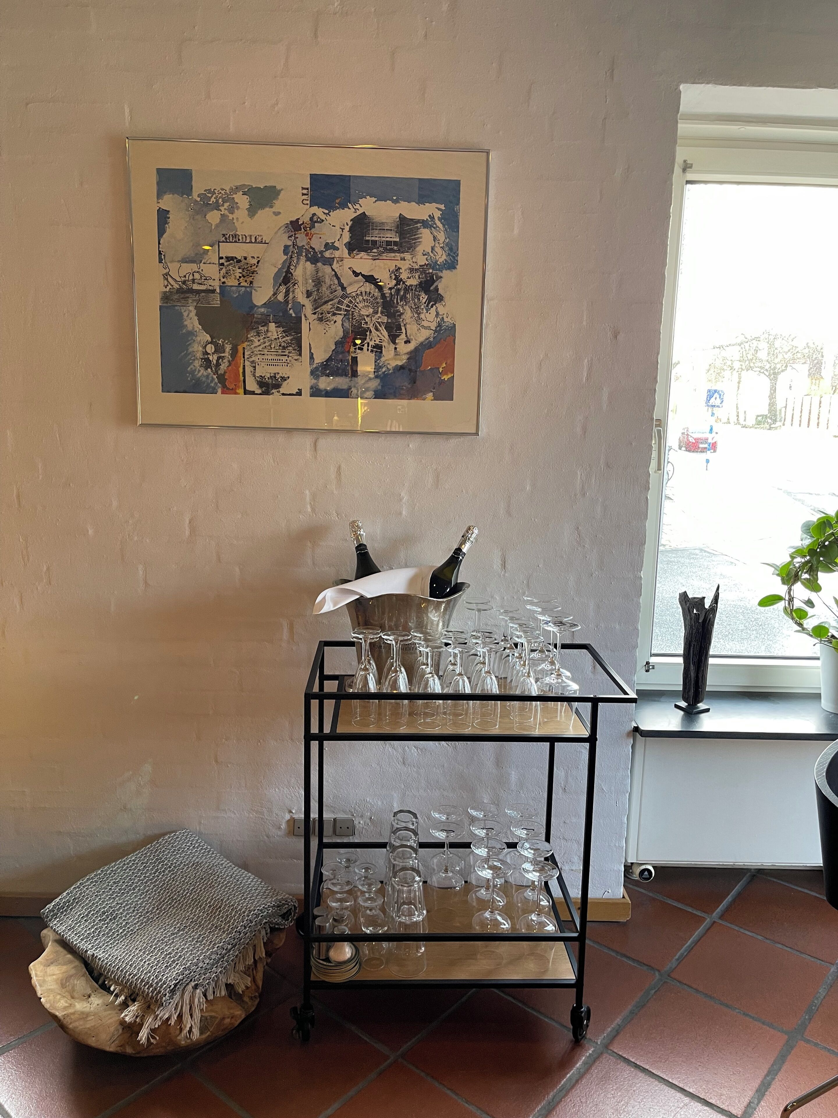 A room with a tiled floor, a window with a view of the street, a framed abstract painting on the wall, and a cart with wine glasses and bottles.