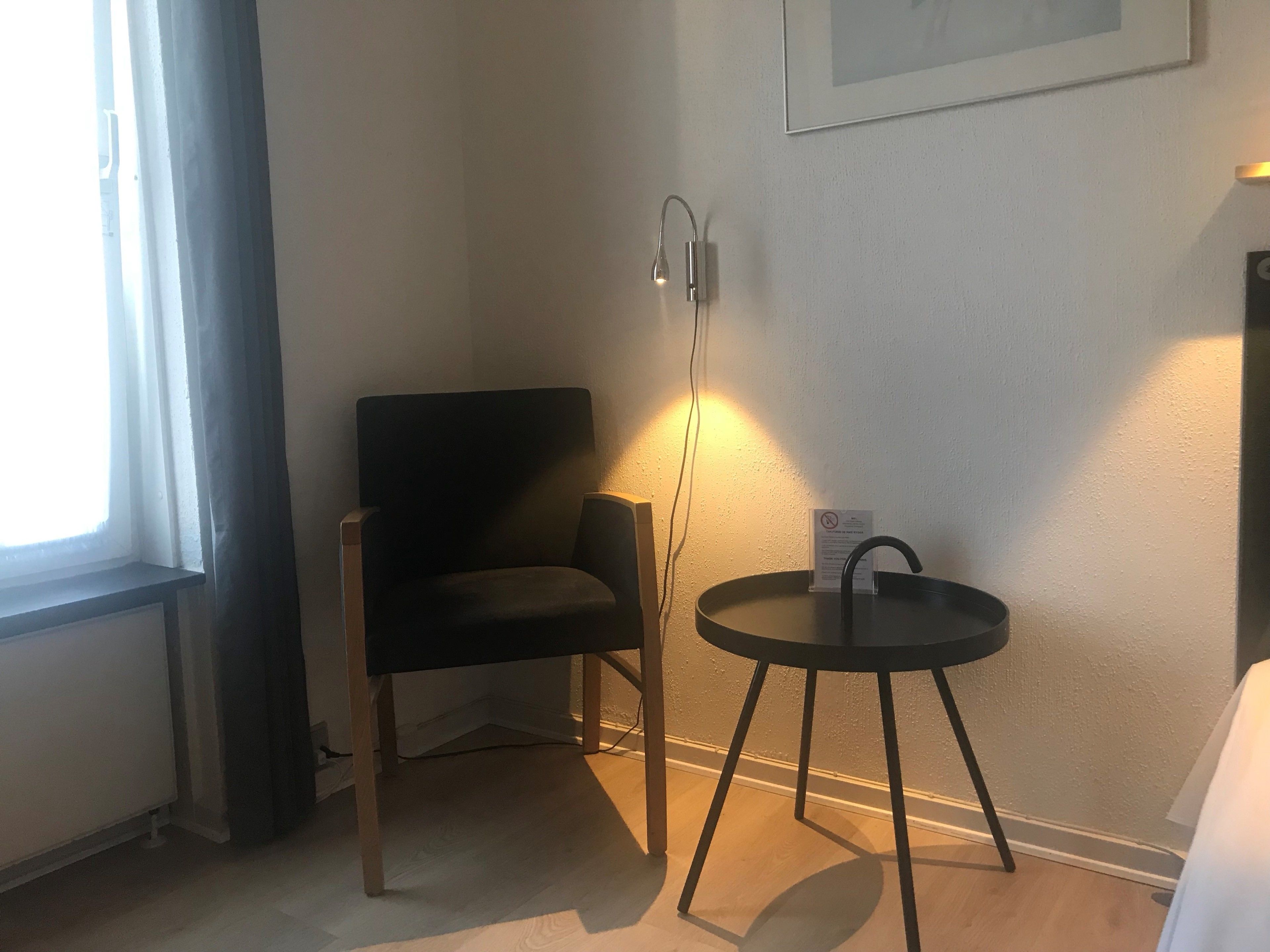 Een hoekzaal met een zwarte stoel, een klein rond tafeltje met een zwart onderstel, een wandlamp en een ingelijst kunstwerk.