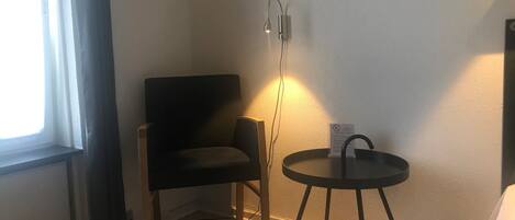 Een hoekzaal met een zwarte stoel, een klein rond tafeltje met een zwart onderstel, een wandlamp en een ingelijst kunstwerk.