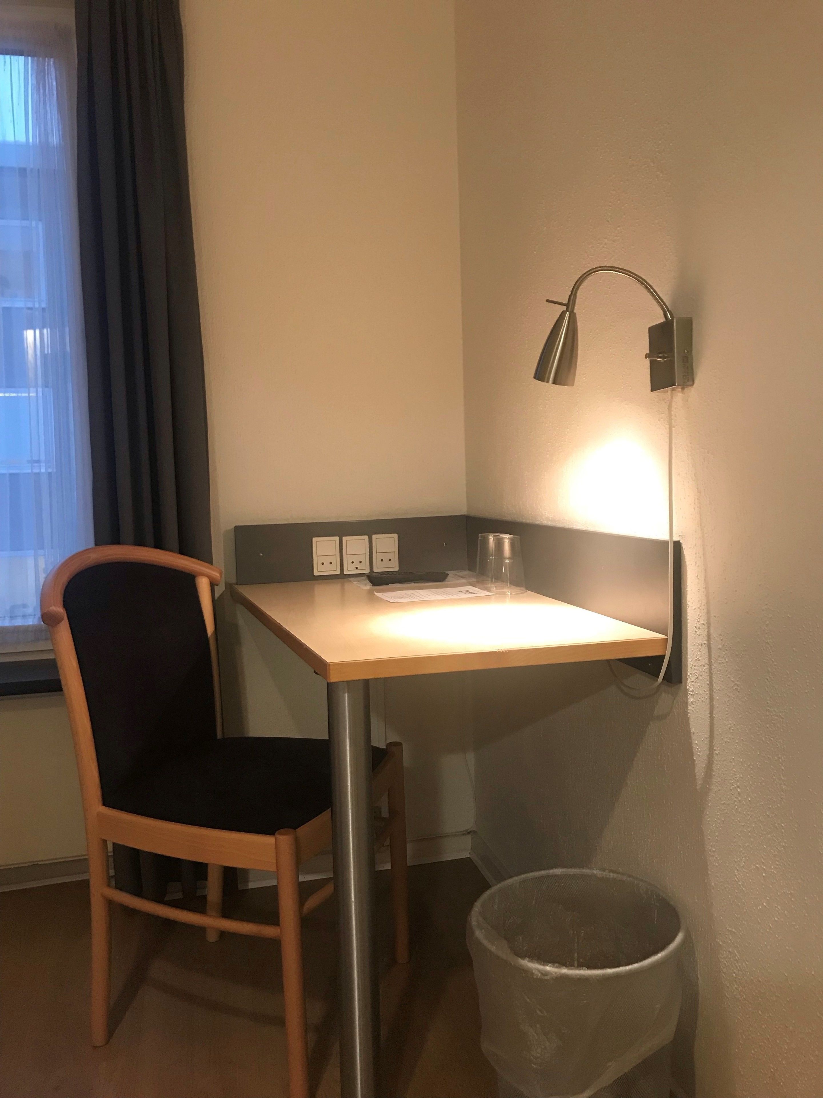A small, well-lit room with a wooden desk, a chair, a wall-mounted lamp, and a trash bin.