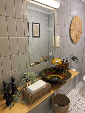 Un baño moderno con un espejo grande, un lavabo original y un estante de madera con artículos de aseo.