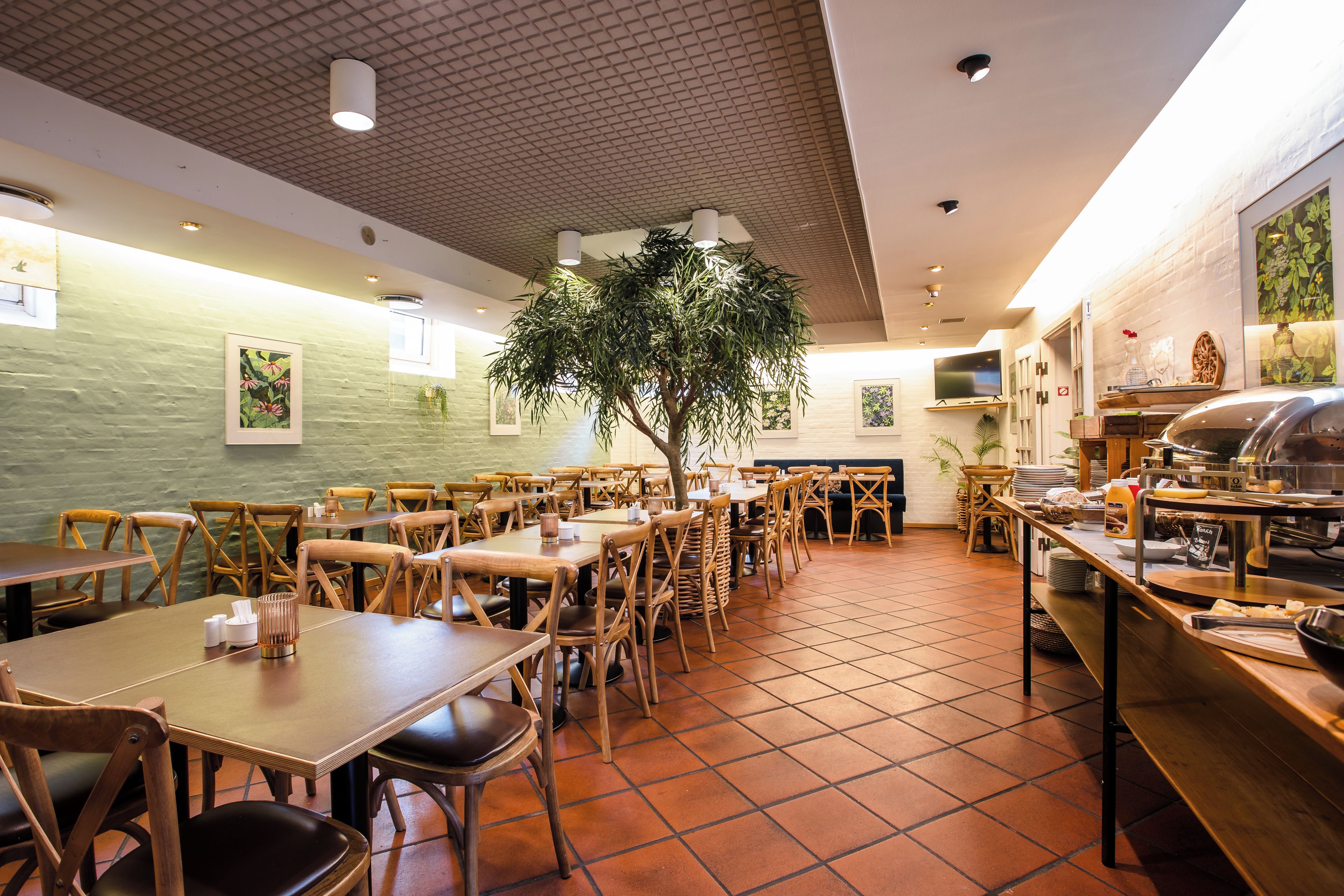 A restaurant interior with wooden tables and chairs, a large potted tree, and a buffet area.