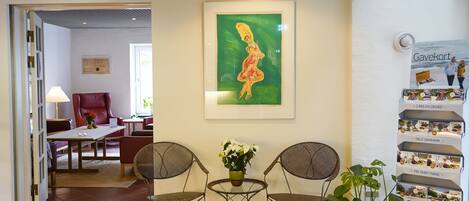 A reception area with a framed painting, two chairs, a small table with a vase of flowers, and a display of food boxes.
