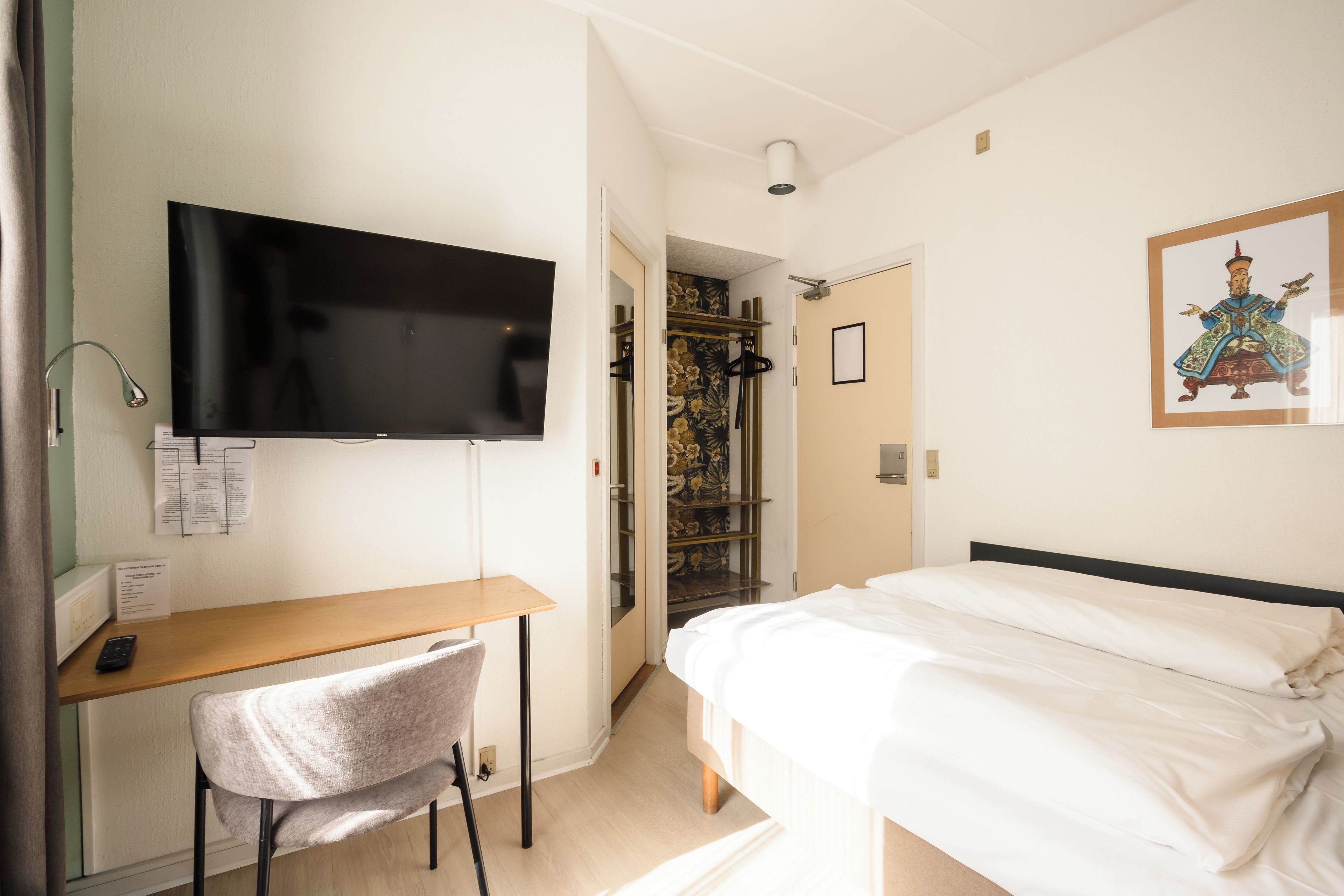 A hotel room with a bed, a desk, a chair, a television, a wardrobe, and a painting on the wall.