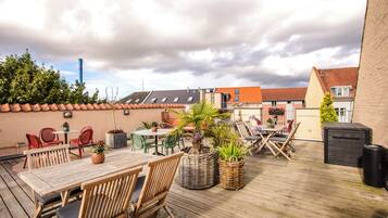 Eine Holzterrasse mit Gartenmöbeln, Blumentöpfen und Blick auf Gebäude im Hintergrund.