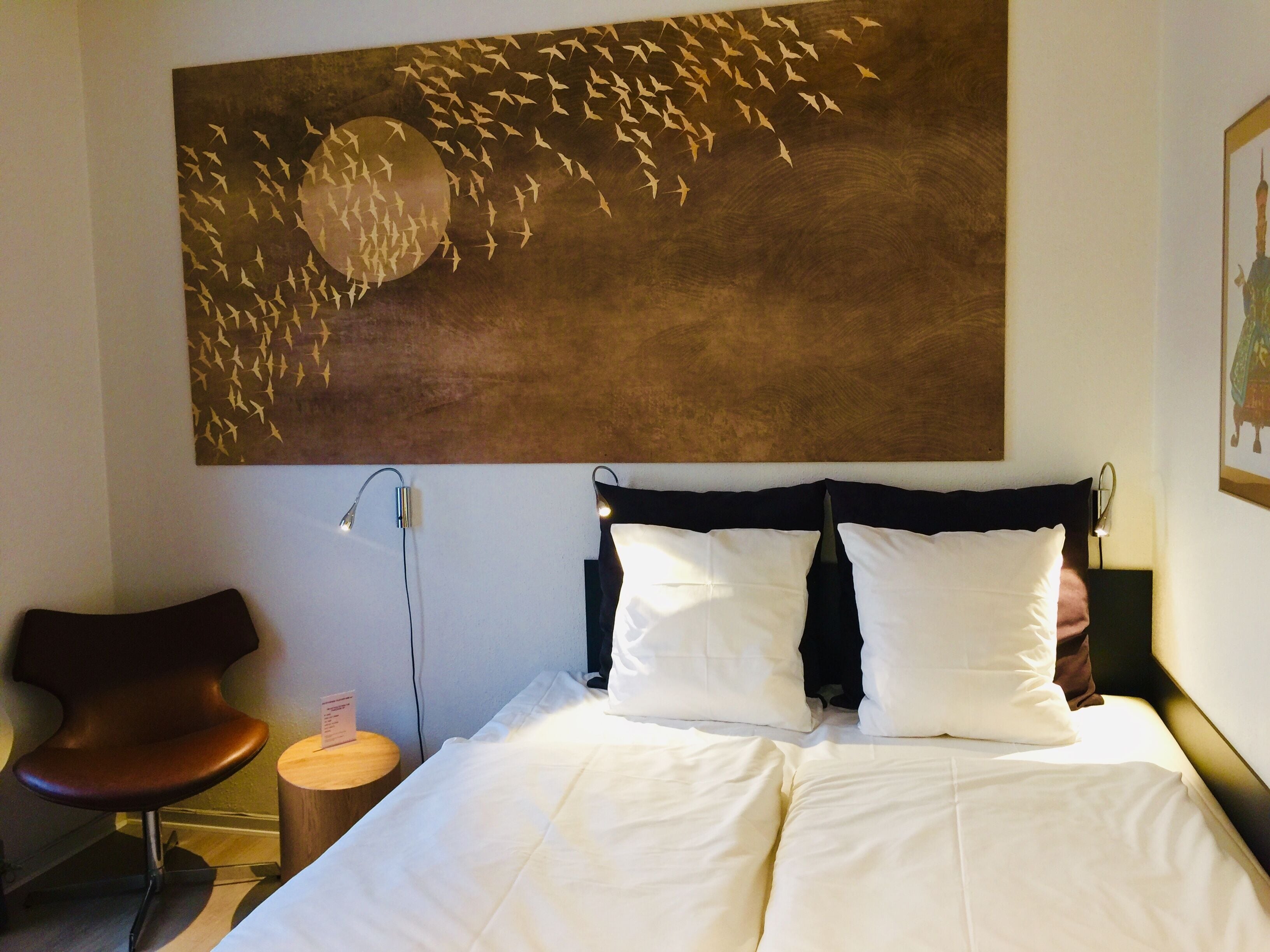 A hotel room with a large abstract painting, a bed with white linens, a wooden nightstand, a modern chair, and a wall-mounted lamp.