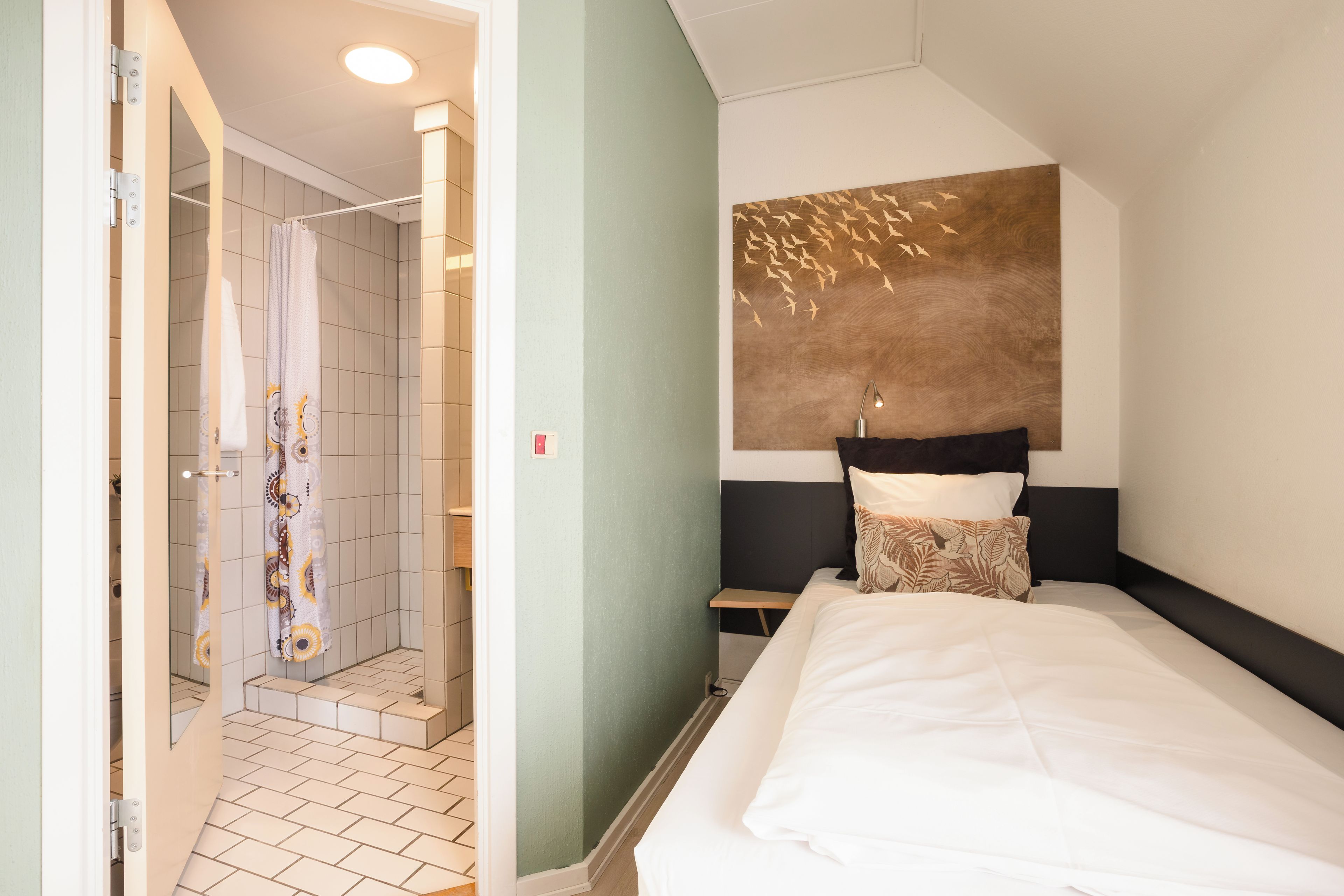 Une chambre d’hôtel avec une douche, un lit avec du linge de lit blanc, une tête de lit ornée d’un panneau décoratif et une table de chevet avec une lampe.