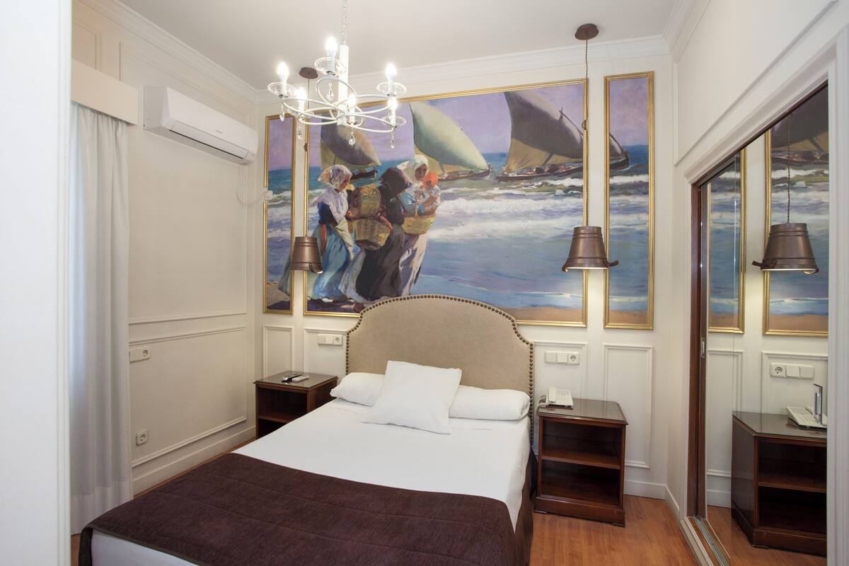 Un cuarto de hotel con una cama grande, una araña, un cuadro de una escena náutica y vistas al mar a través de una ventana.