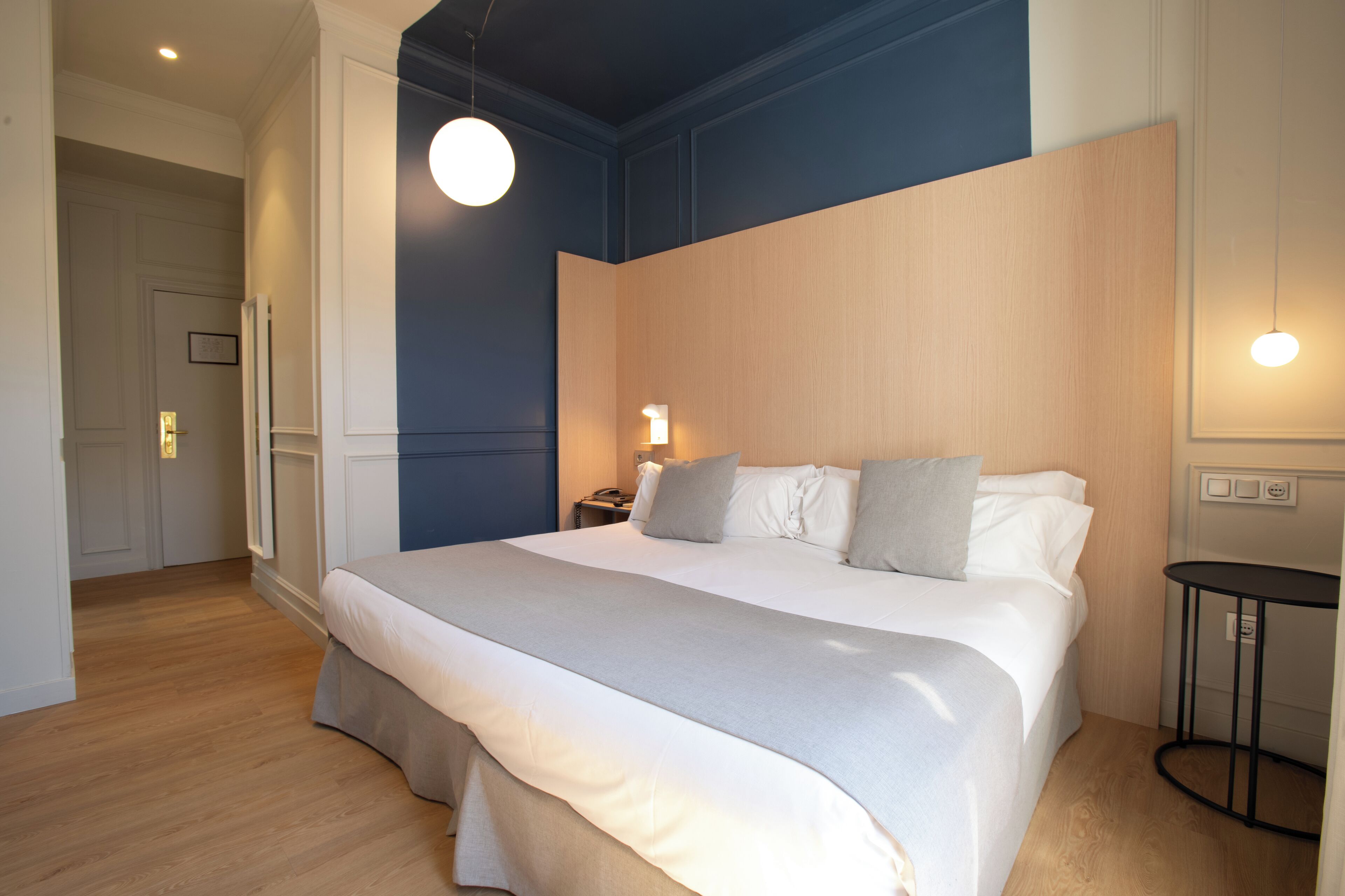 A neatly made bed with a white comforter and gray throw, a bedside lamp, and a wooden headboard.