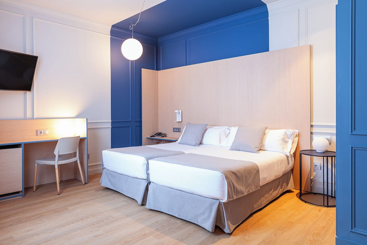 Ein Doppelbett mit Holzfußteil, ein Schreibtisch mit Stuhl, ein runder Tisch und ein an der Wand befestigter Fernseher.