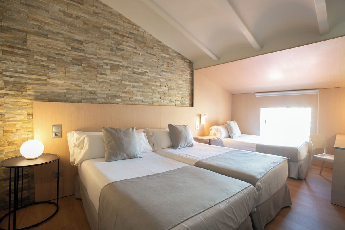 Ein Hotelzimmer mit zwei Betten, einer Steinwand, einem runden Tisch und einem Schreibtisch mit einem Stuhl.