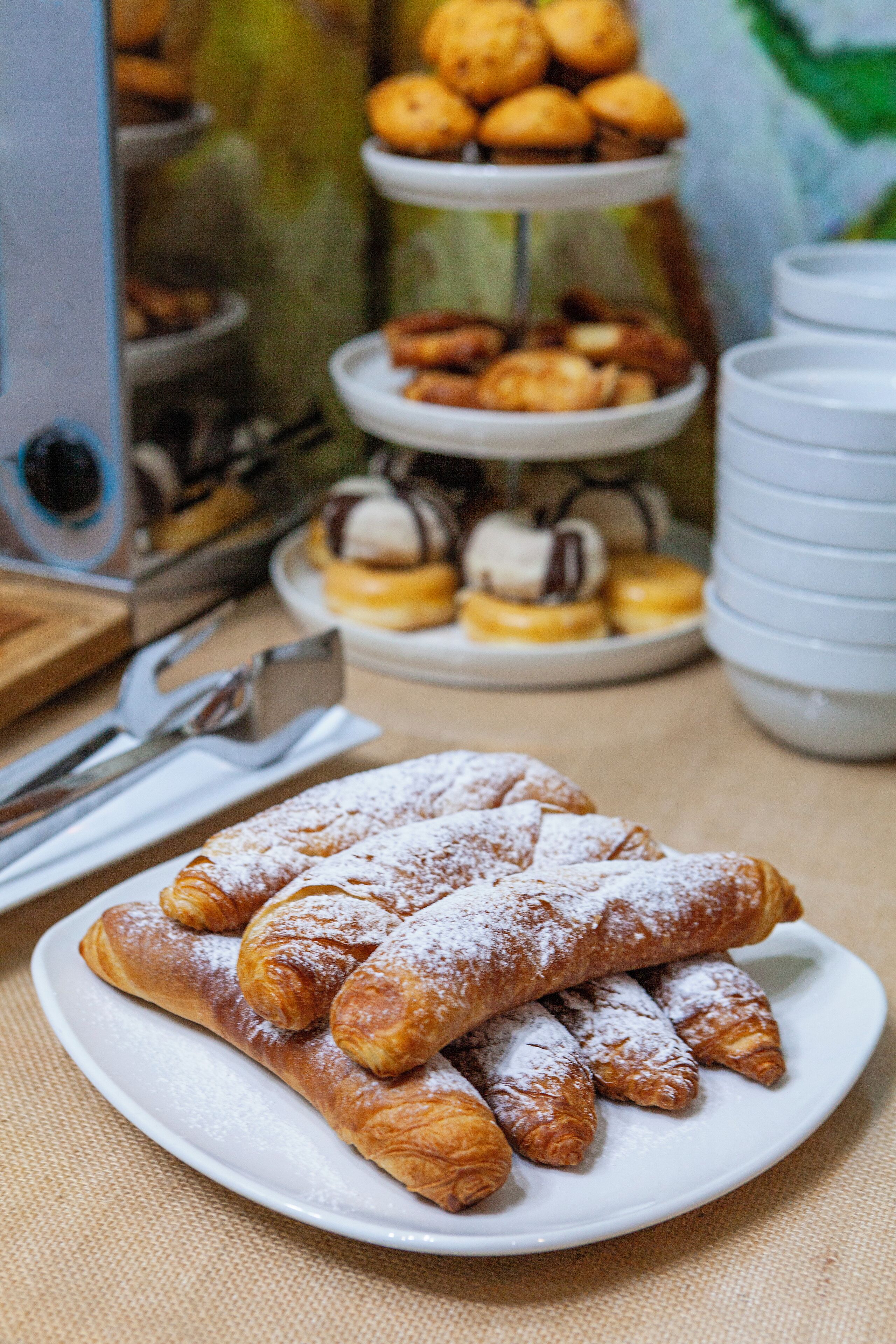 A plate of pastries with powdered sugar, a tiered display of various baked goods, and stacks of white plates.
