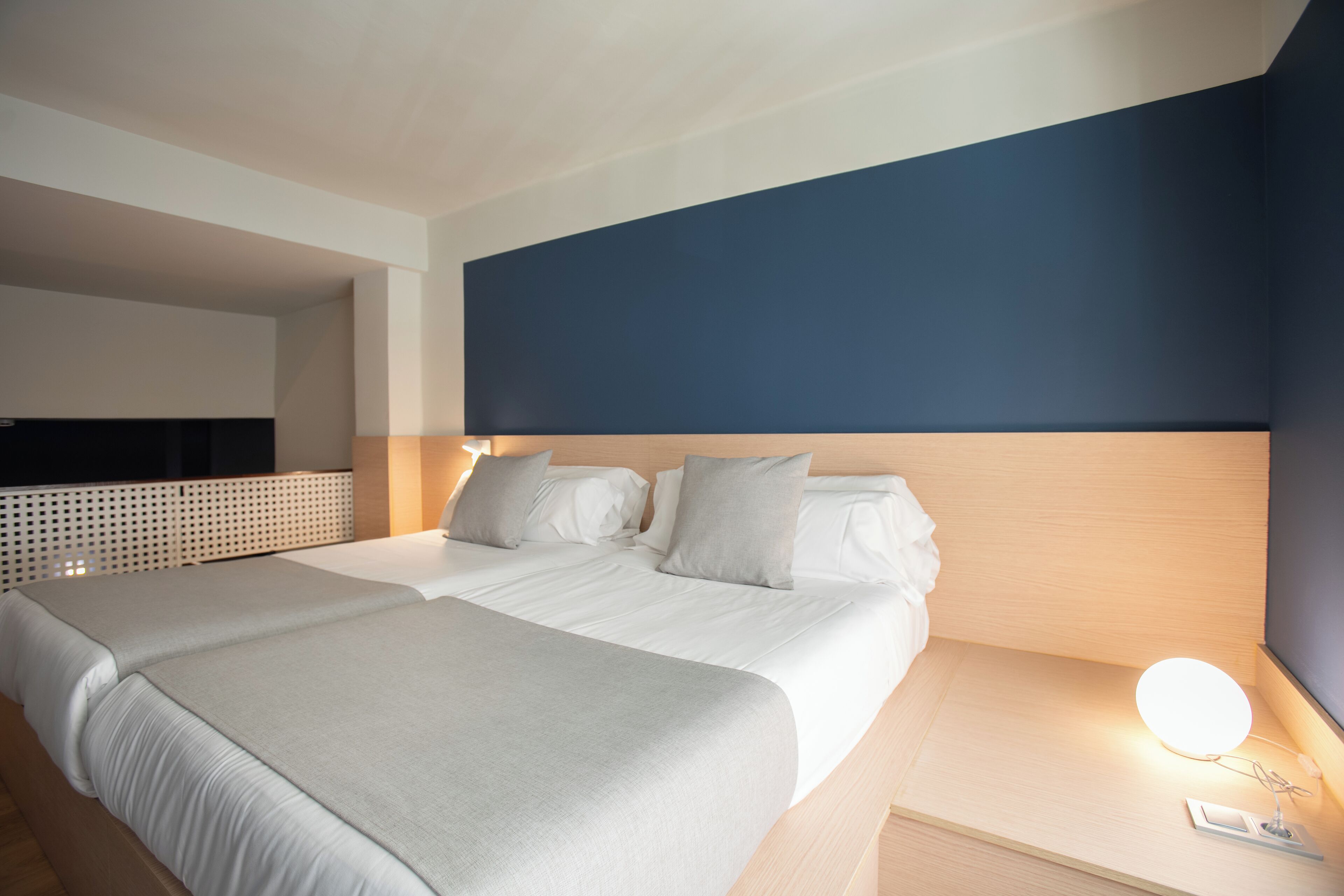 Ein Doppelbett mit weißen Leinen und einer grauen Tagesdecke, ein Nachttisch mit Lampe und eine dunkelblaue Akzentwand.