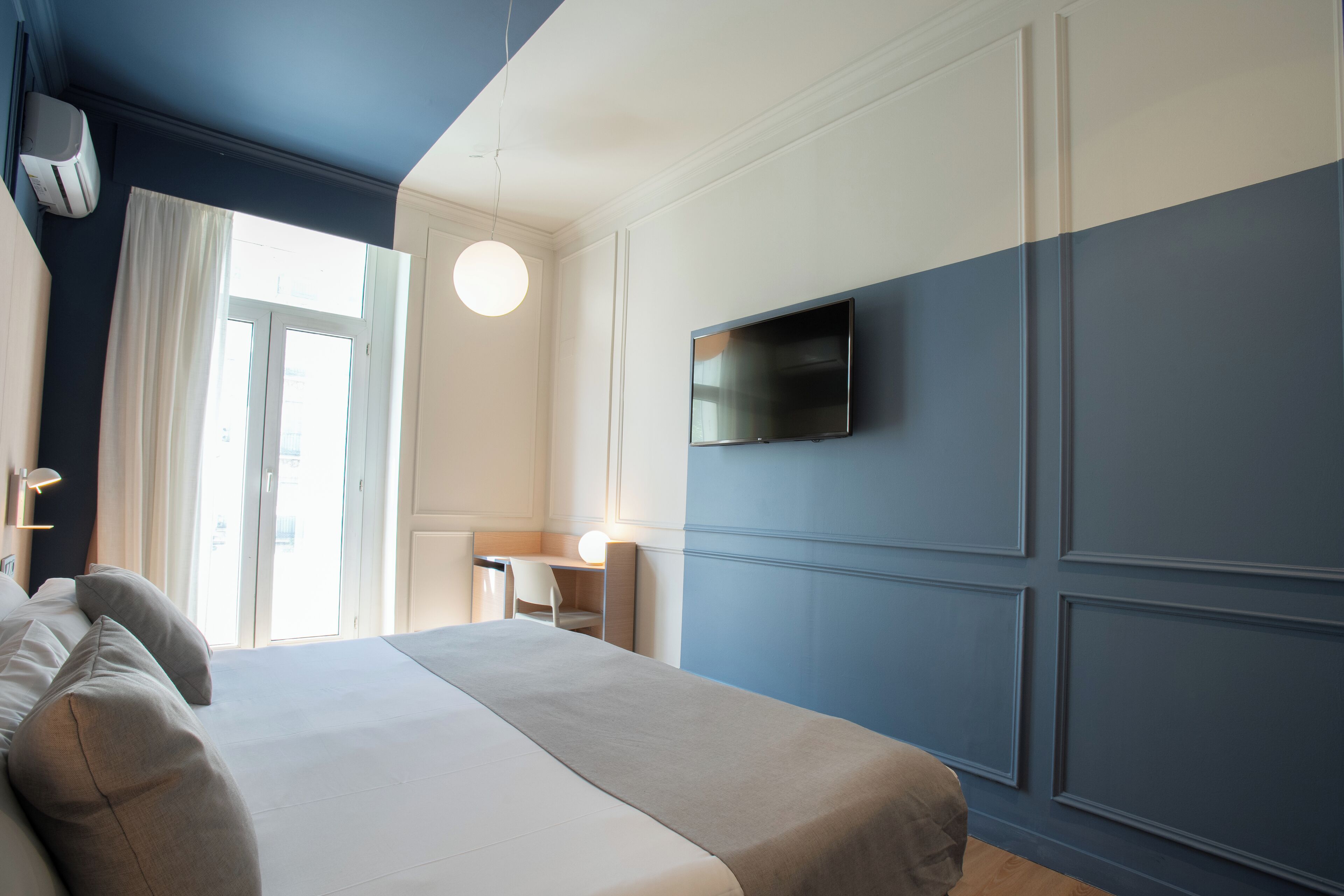 A modern hotel room with a large bed, a desk, a chair, and a flat-screen TV mounted on the wall.