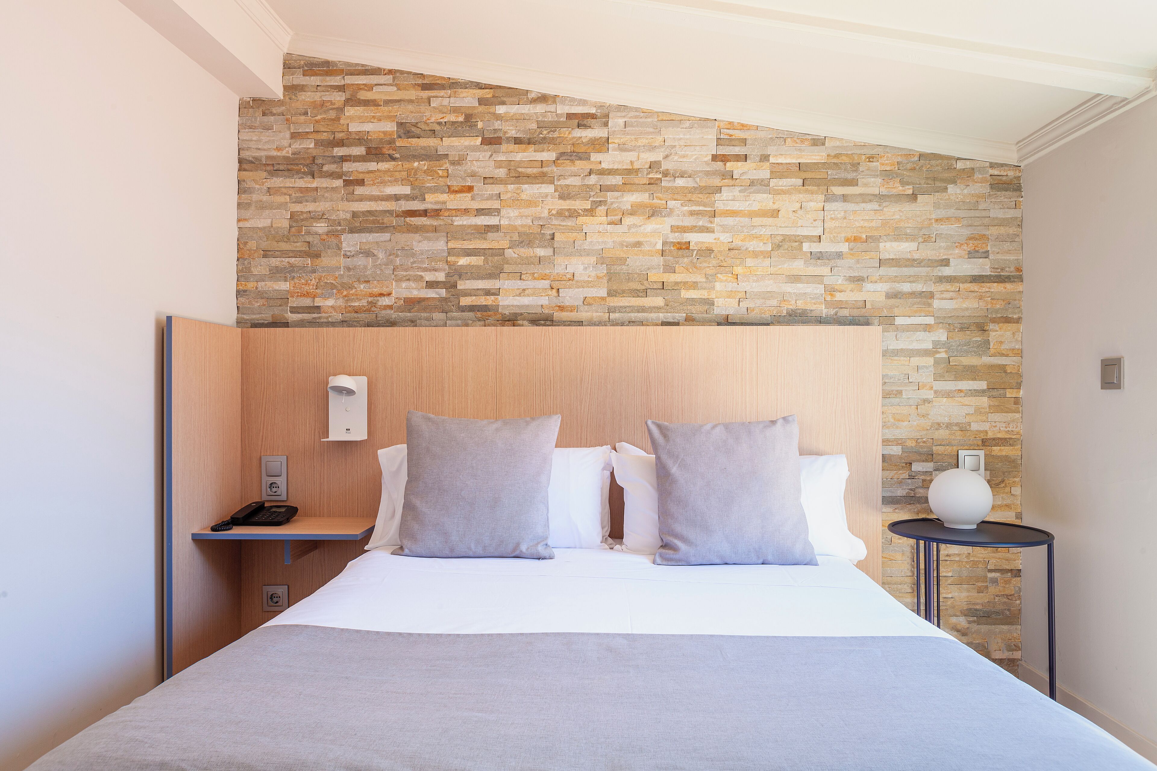 Hotellihuone, jossa on kiviseinä, puinen sängynpääty, harmailla tekstiileillä varustettu sänky, yöpöytä, jossa on puhelin, ja pieni pöytä, jossa on maljakko.