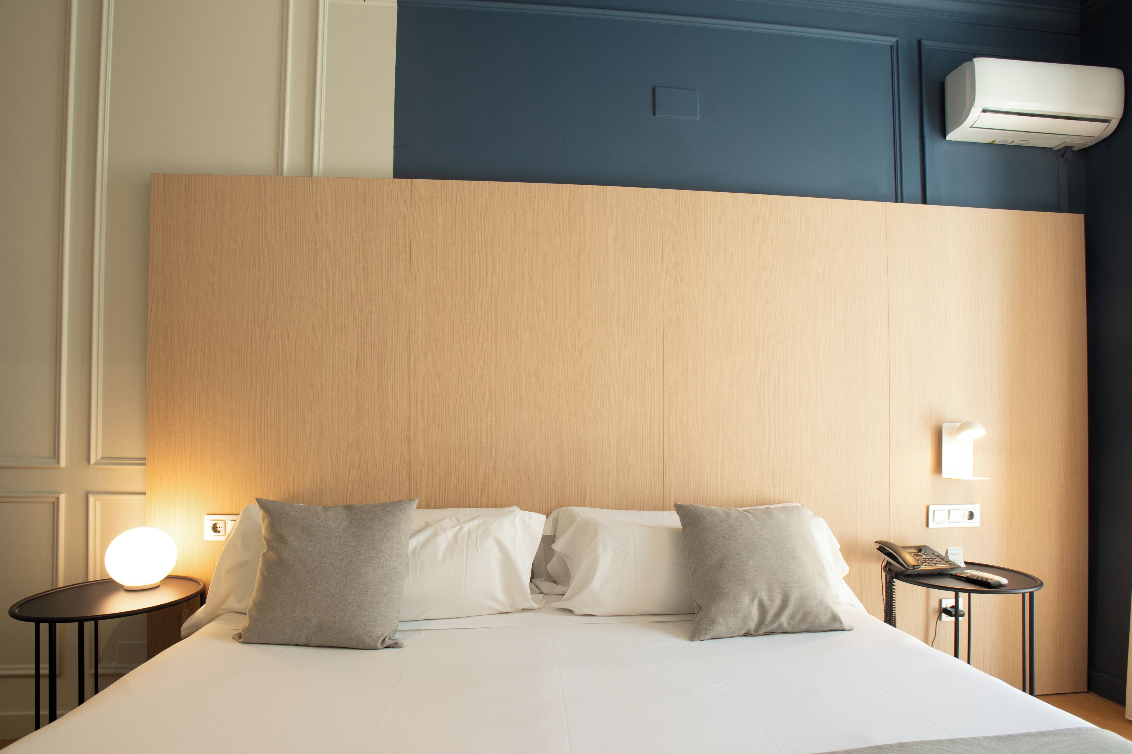 Ein Hotelzimmer mit einem hölzernen Kopfteil, einem Bett mit weißer Bettwäsche, zwei Nachttischen, einem an der Wand befestigten Telefon und einer Klimaanlage.