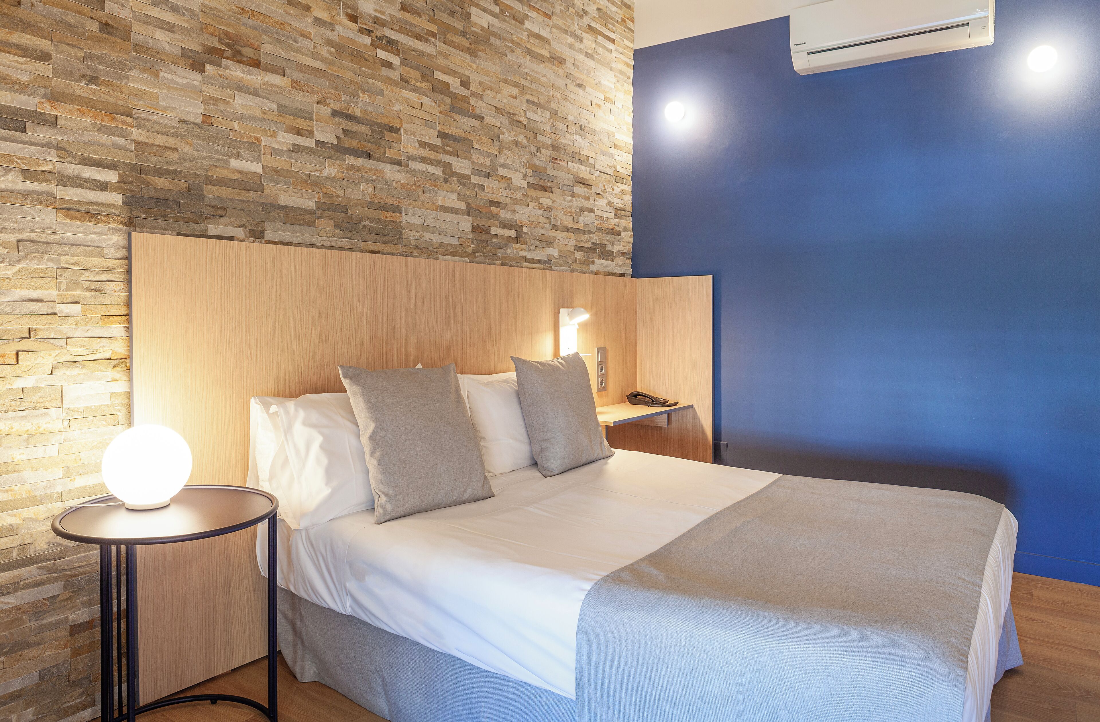 Un lit bien fait, avec une tête de lit, une table de chevet avec une lampe et un mur orné d’un motif texturé.