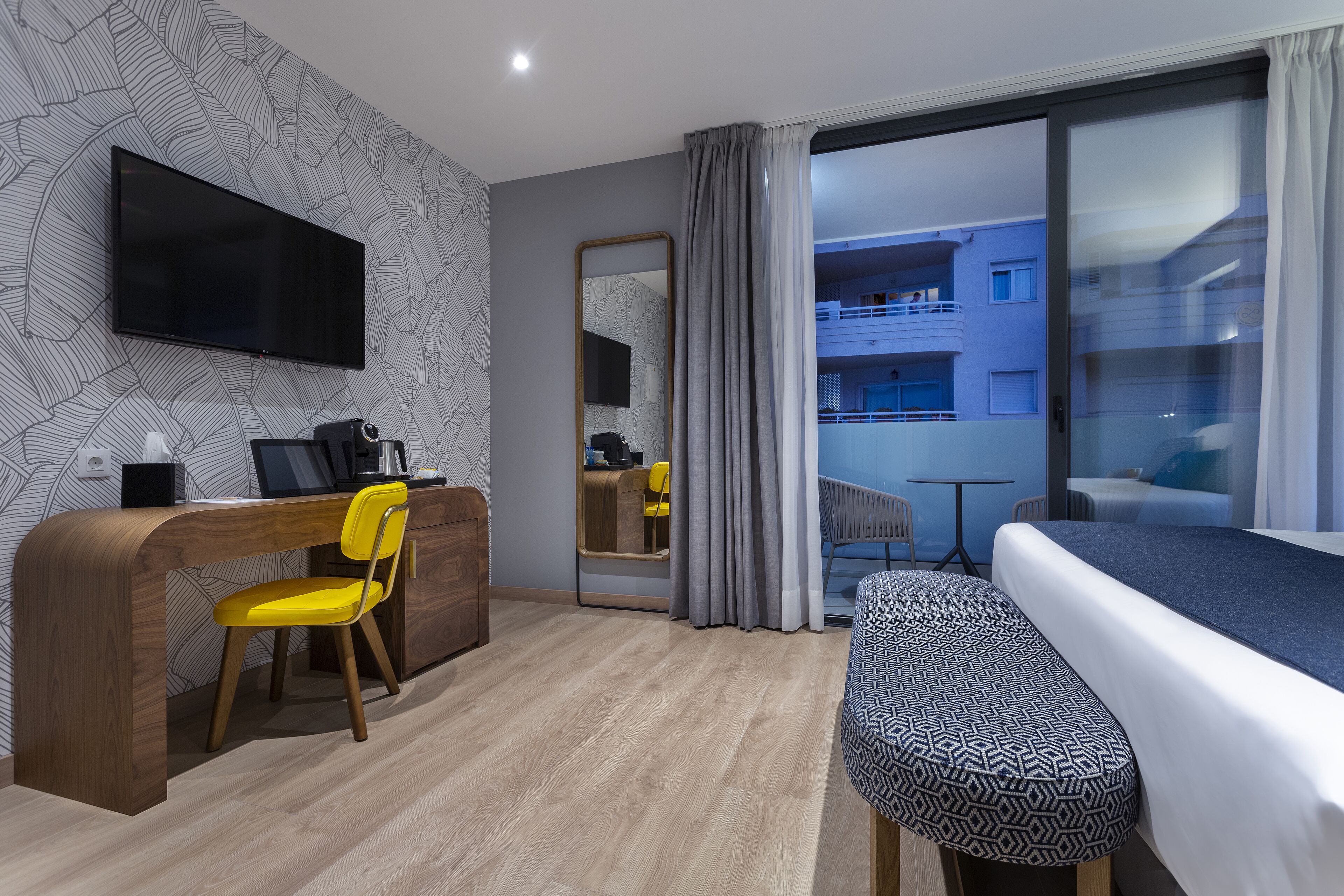 Habitación de hotel moderna con un escritorio de madera, una silla amarilla, una cama con ropa de cama estampada, un espejo grande y vistas del exterior del edificio a través de una puerta de vidrio.