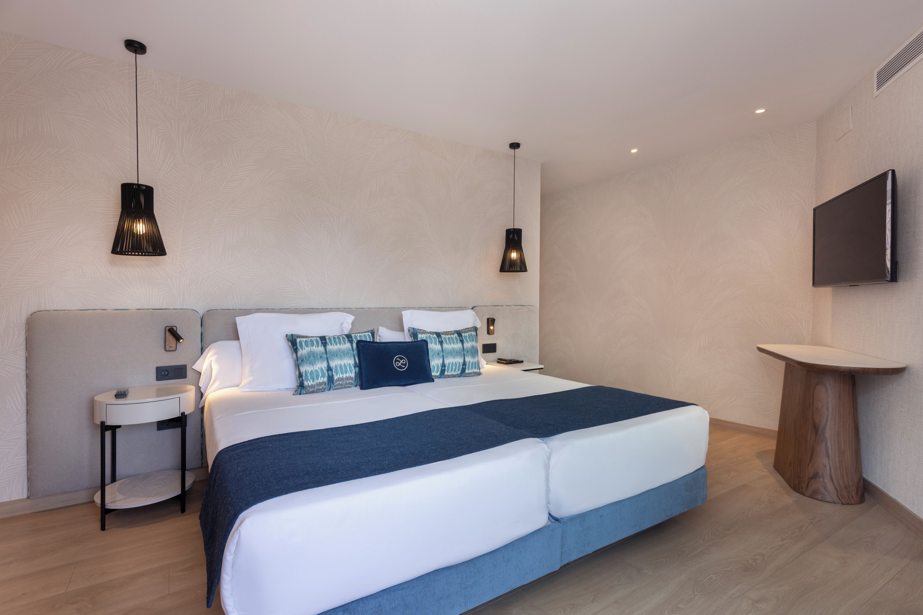 A modern hotel room with a large bed, two bedside tables, a TV, and pendant lighting.