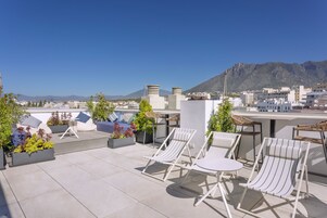 Een dakterras met lounge fauteuils, plantenbakken met groen, en uitzicht op de stad en de bergen.