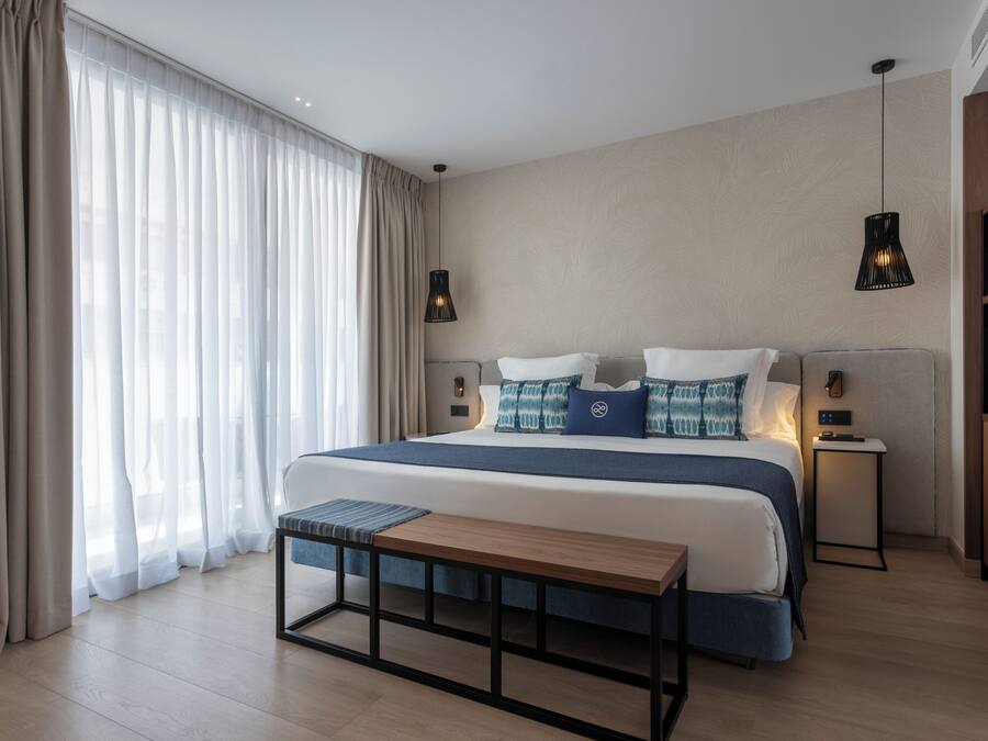 Une chambre à coucher moderne avec un grand lit, des tables de chevet, un banc et une vue sur l’extérieur.