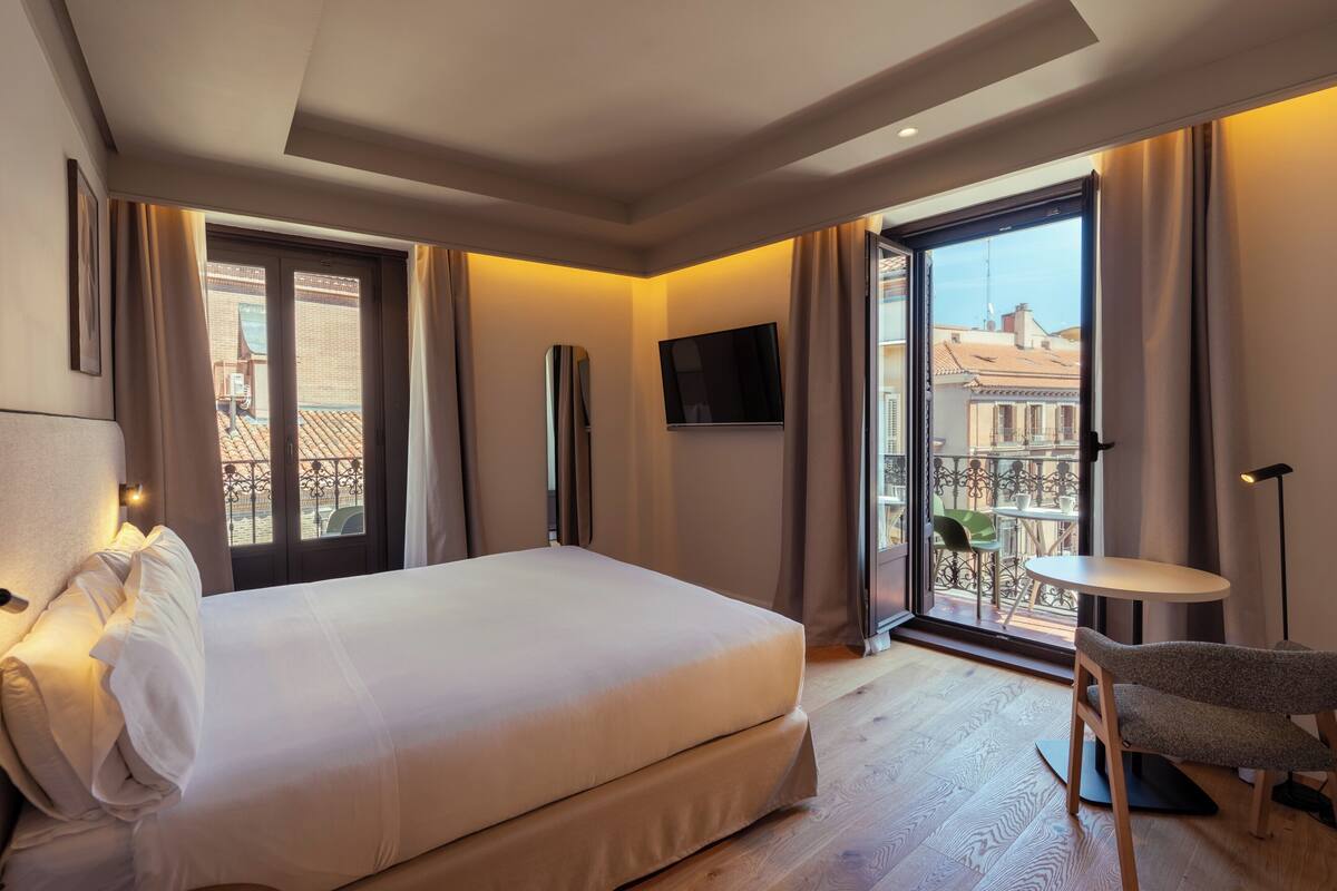 A hotel room with a bed, a TV, a balcony with a view of buildings, and a small table.
