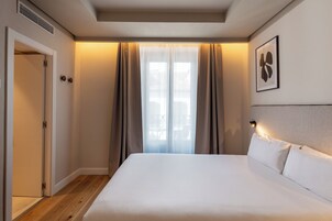 A hotel room with a bed, a door, a window with curtains, and a wall-mounted light fixture.