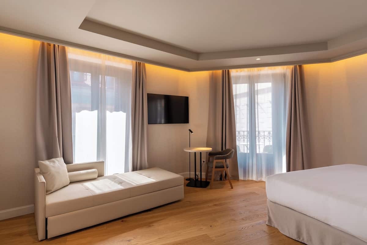 A modern hotel room with a sofa, a small table with a chair, a bed, and a TV mounted on the wall.