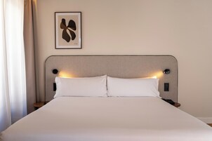 Una cama bien hecha con ropa blanca, un cabecero con iluminación integrada, una obra de arte abstracta enmarcada en la pared y una mesita de noche con un teléfono.