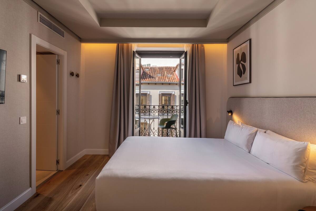 A hotel room with a bed, a view of a balcony with a chair, a door, and a wall-mounted light fixture.