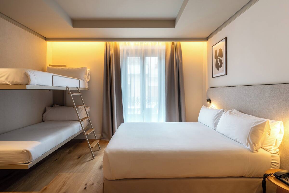 A hotel room with a bunk bed, a single bed, a bedside table with a lamp, and a window with curtains.