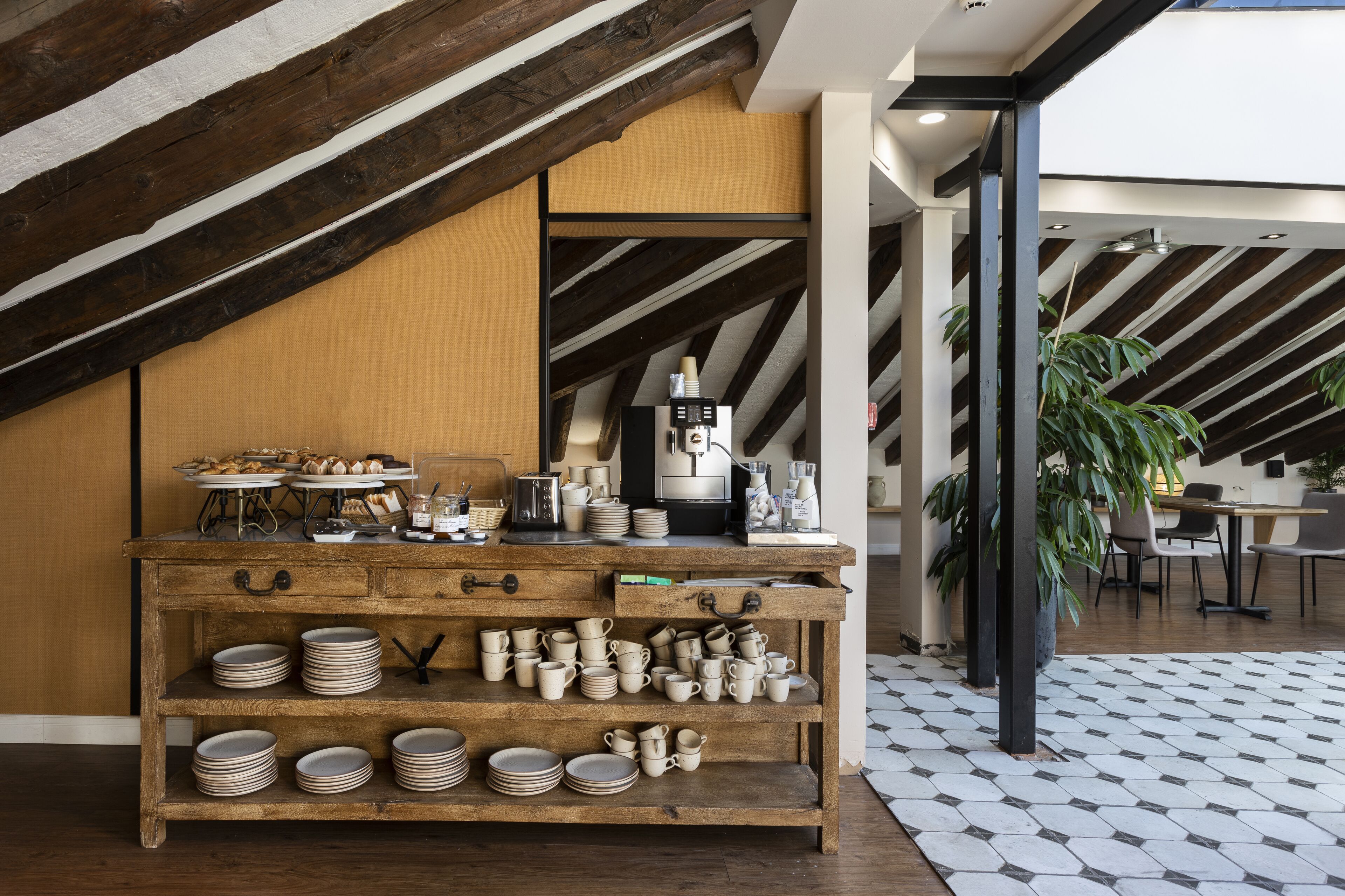 Una mesa de madera rústica con platos y tazas, una cafetera y una planta en un interior moderno con vigas a la vista y suelo de baldosas.