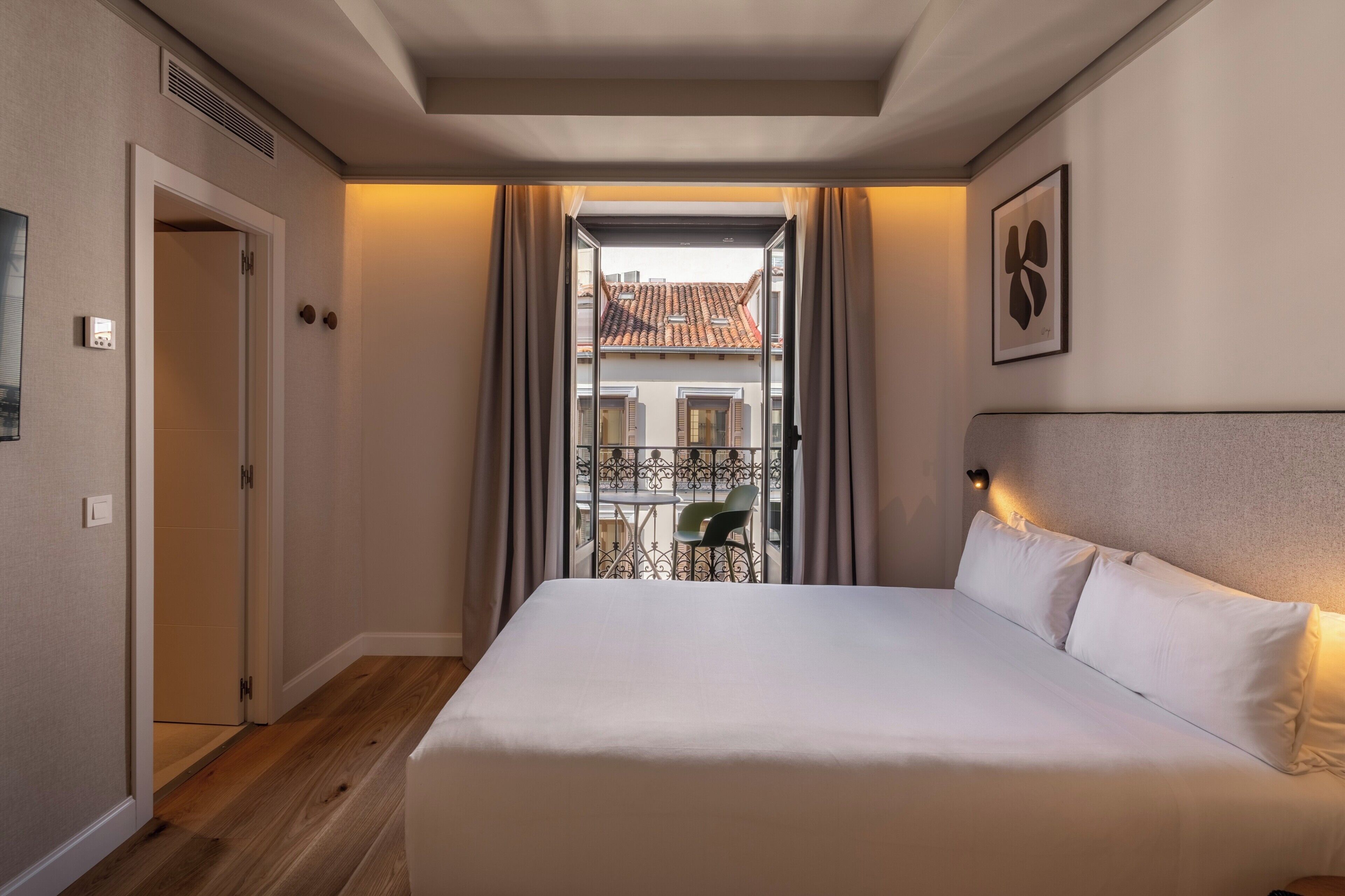 A hotel room with a bed, a view of a balcony with a chair, a door, and a wall-mounted light fixture.