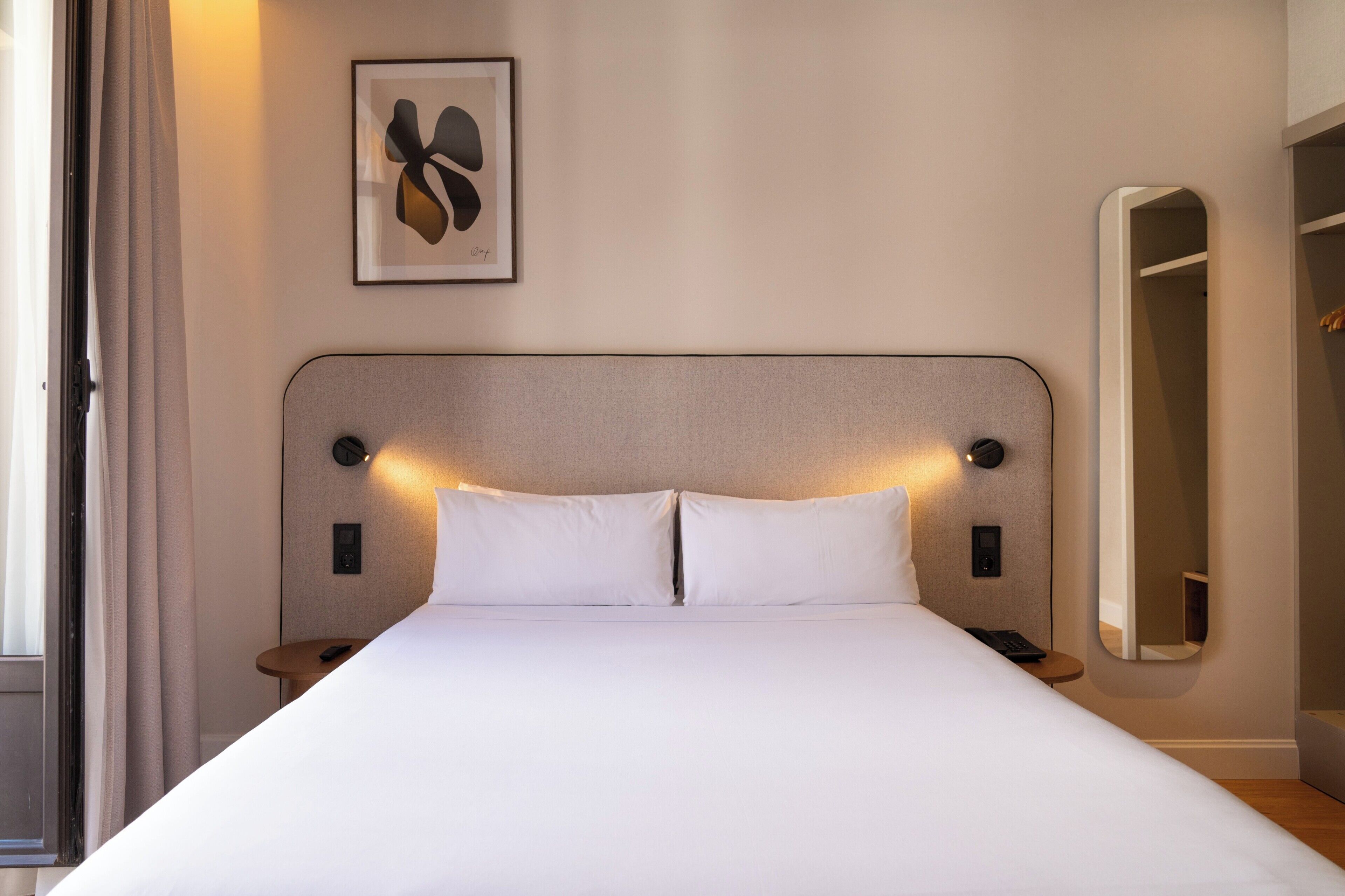 Habitación de hotel con una cama grande, mesitas de noche, un reloj de pared, una obra de arte enmarco y un espejo.
