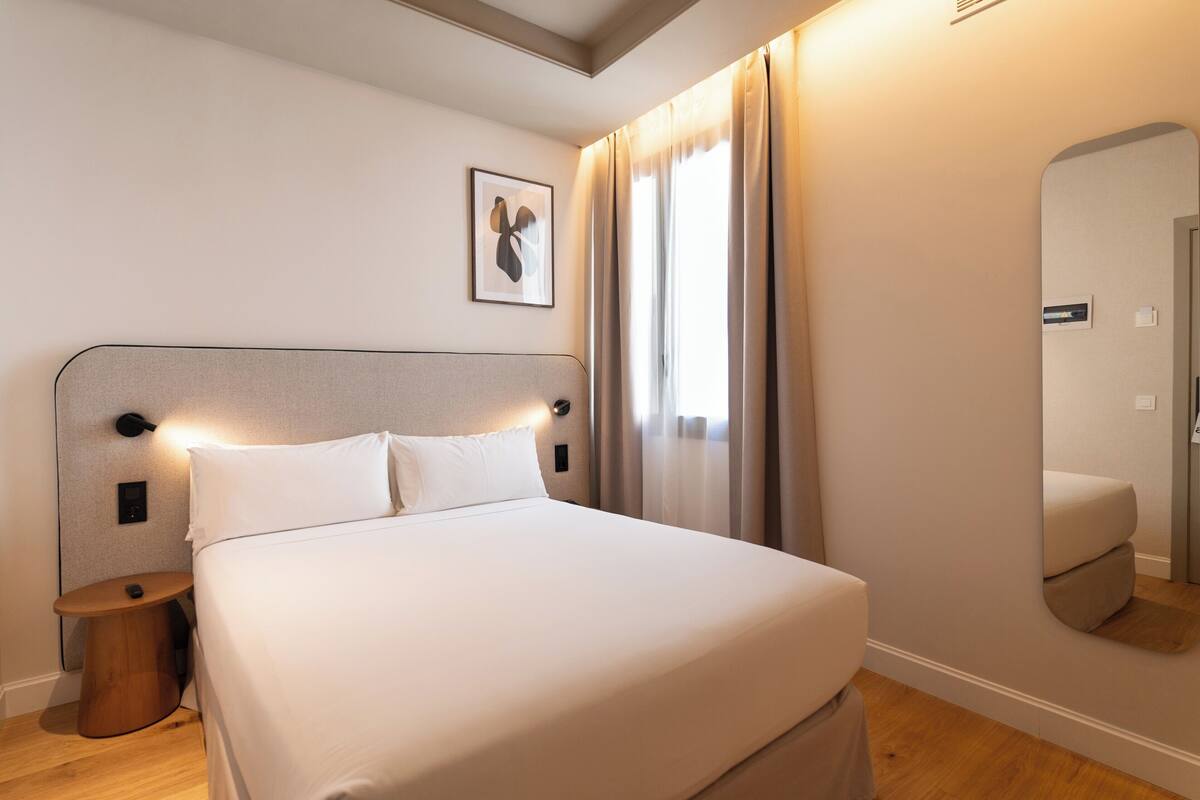 A hotel room with a bed, bedside table, lamp, and a mirror.