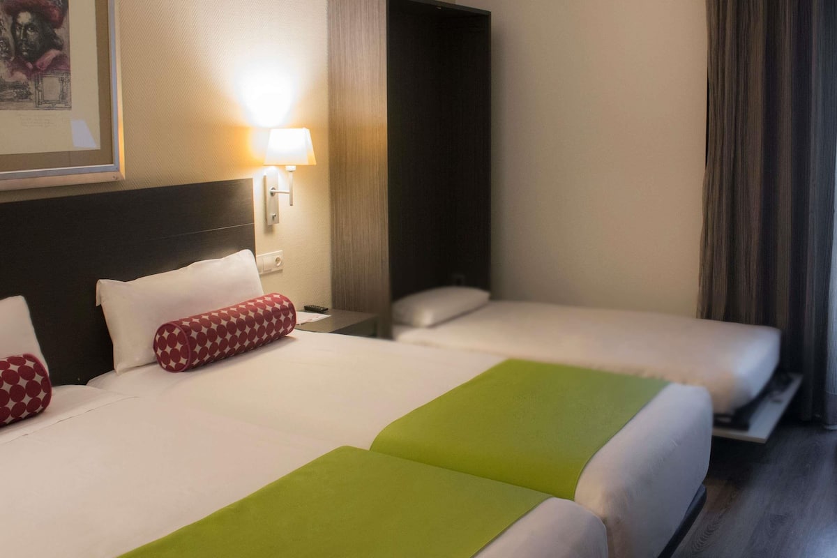 A hotel room with two beds, a nightstand, a lamp, and a painting on the wall.