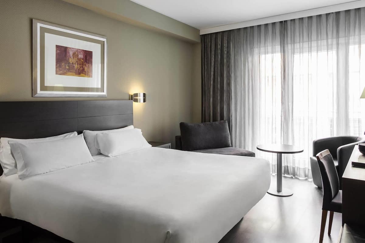 A hotel room with a large bed, a desk, a chair, a small table, and a painting on the wall.