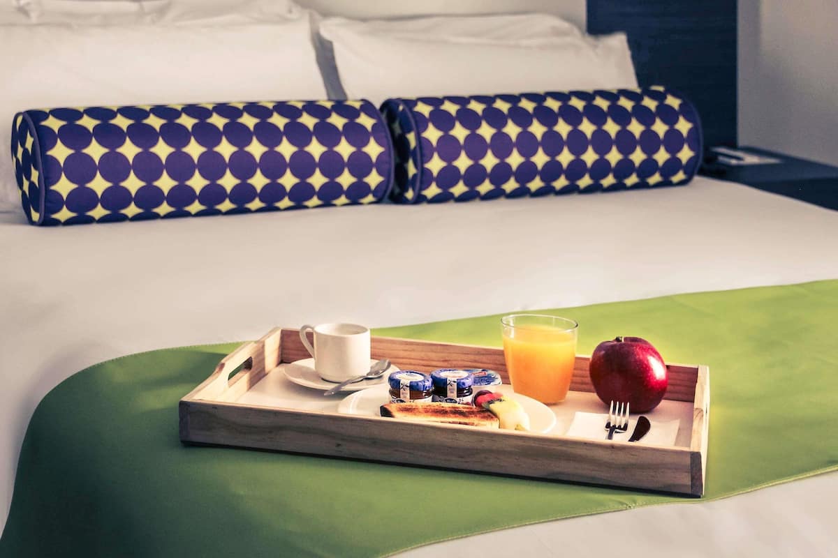 A neatly made breakfast tray with a cup, juice, a banana, and an apple on a bed with a green bedspread and blue patterned pillows.