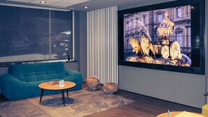 A modern room with a blue sofa, a round wooden table, a large TV screen displaying a statue, and a window with a view of buildings. - Mercure Madrid Centro (Madrid)