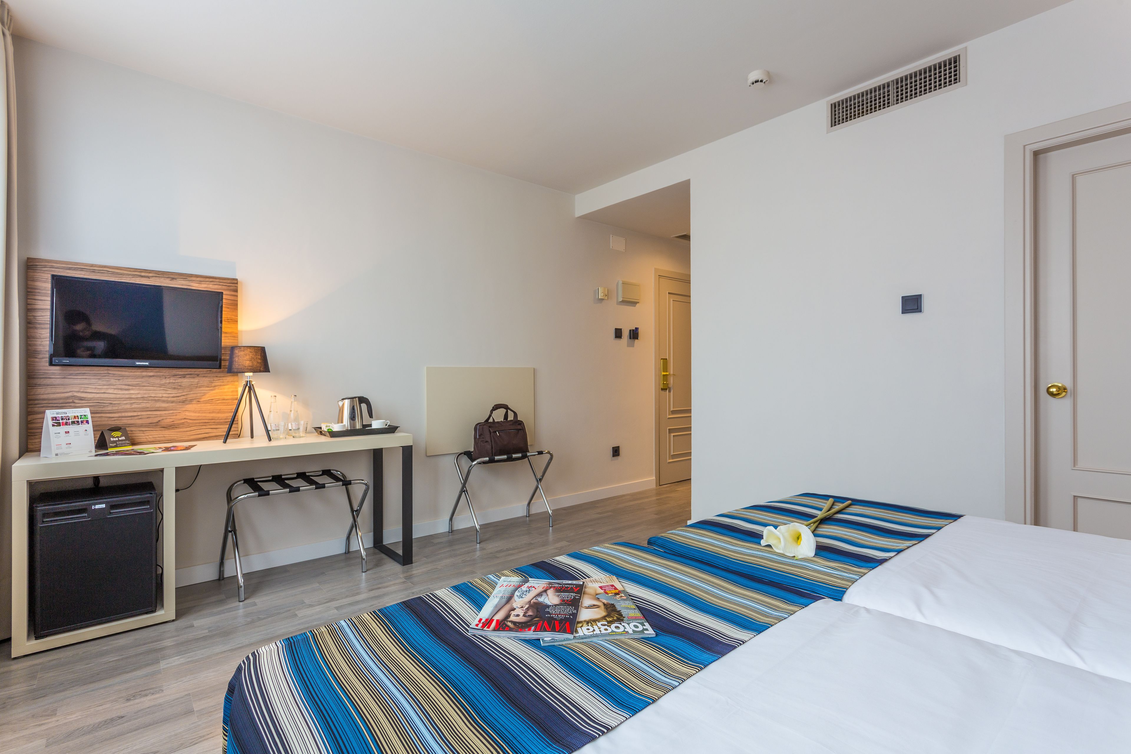 Habitación de hotel con cama, escritorio con televisor, kitchenette pequeña y baño visible a través de una puerta abierta.