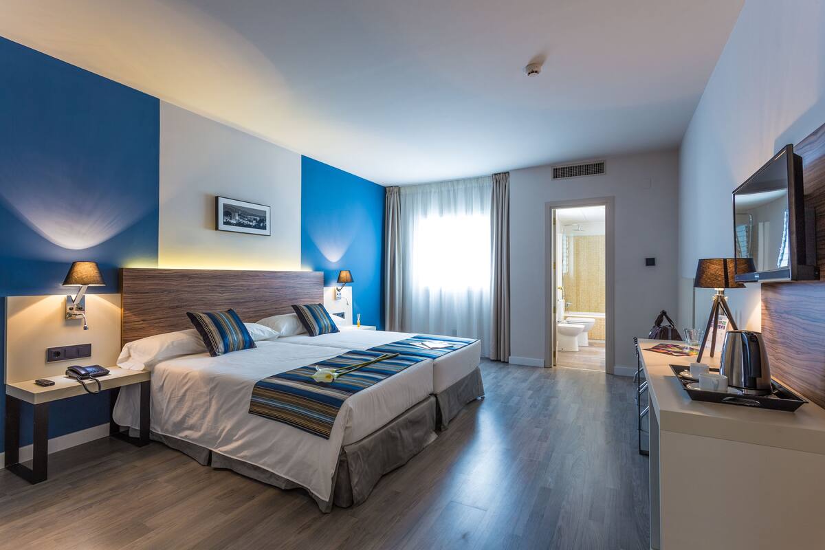 A modern hotel room with a large bed, bedside tables, a desk, and a bathroom visible through an open door.