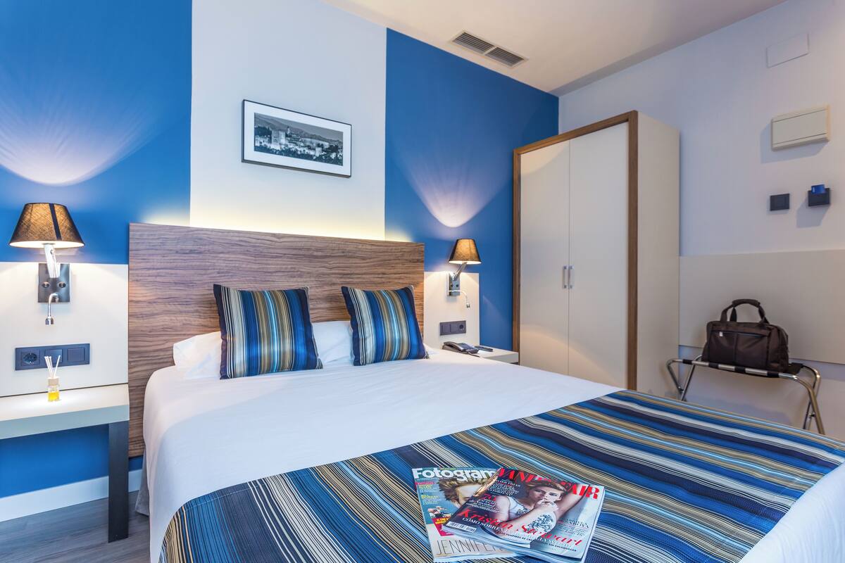 A hotel room with a bed, two bedside lamps, a nightstand, a framed picture on the wall, and a magazine on the bed.