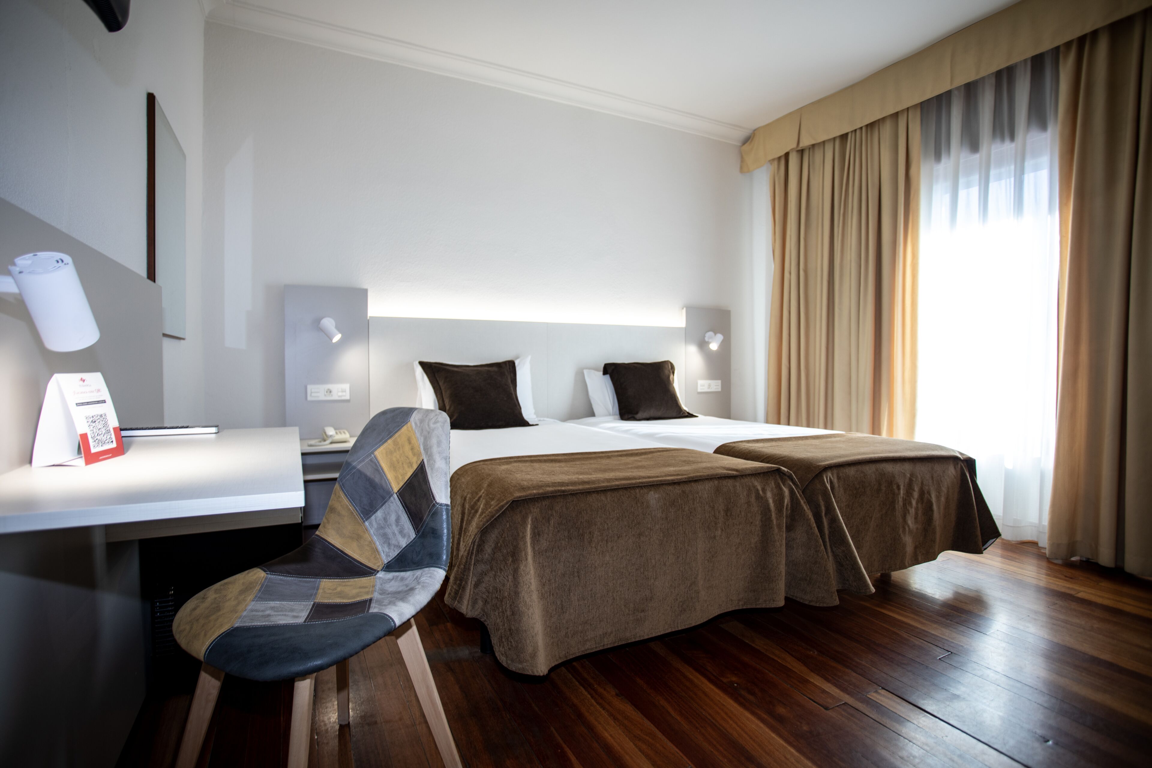 A hotel room with a bed, two bedside tables, a desk, a chair, and a window with curtains.