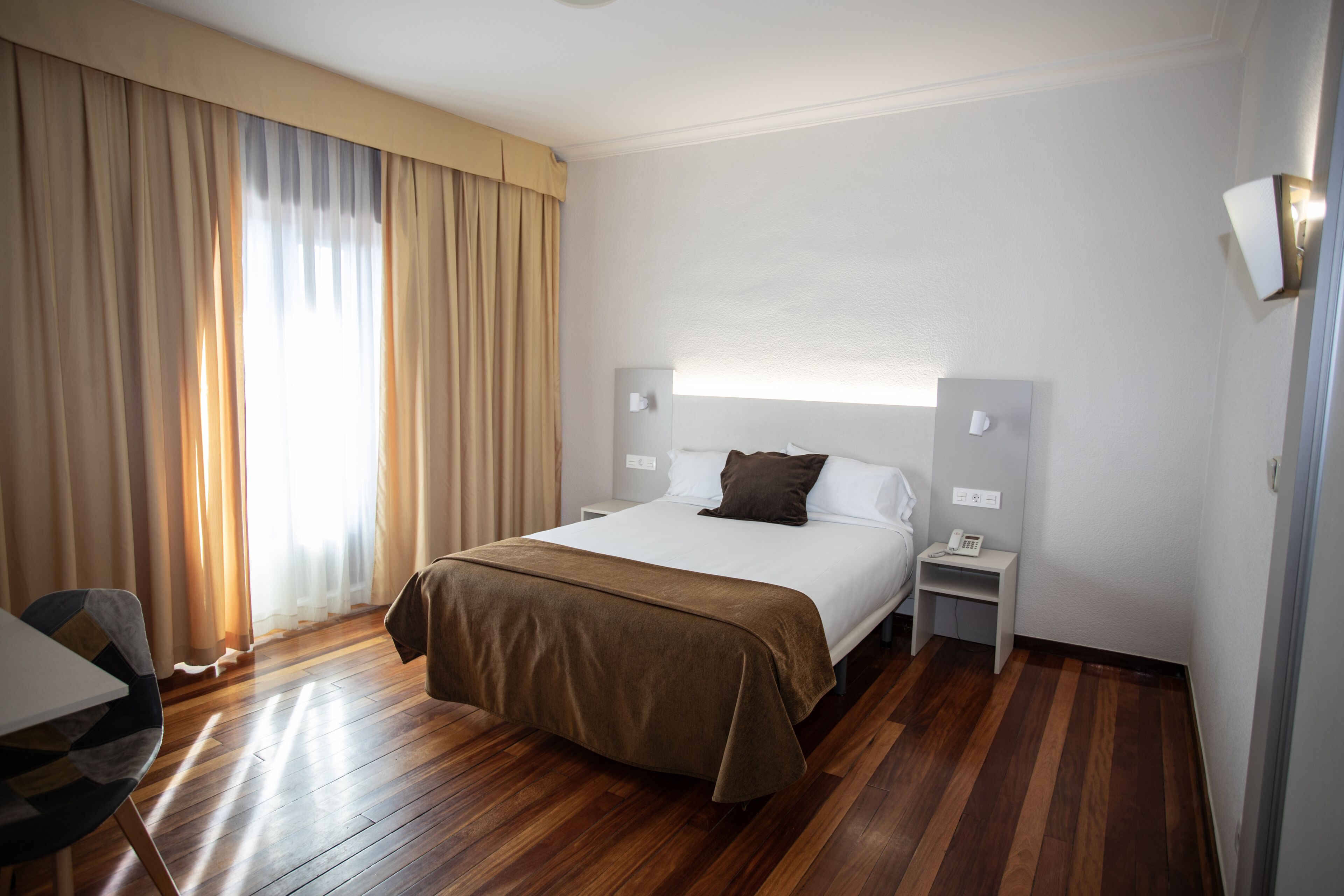 A hotel room with a bed, bedside tables, a chair, and a large window with curtains.