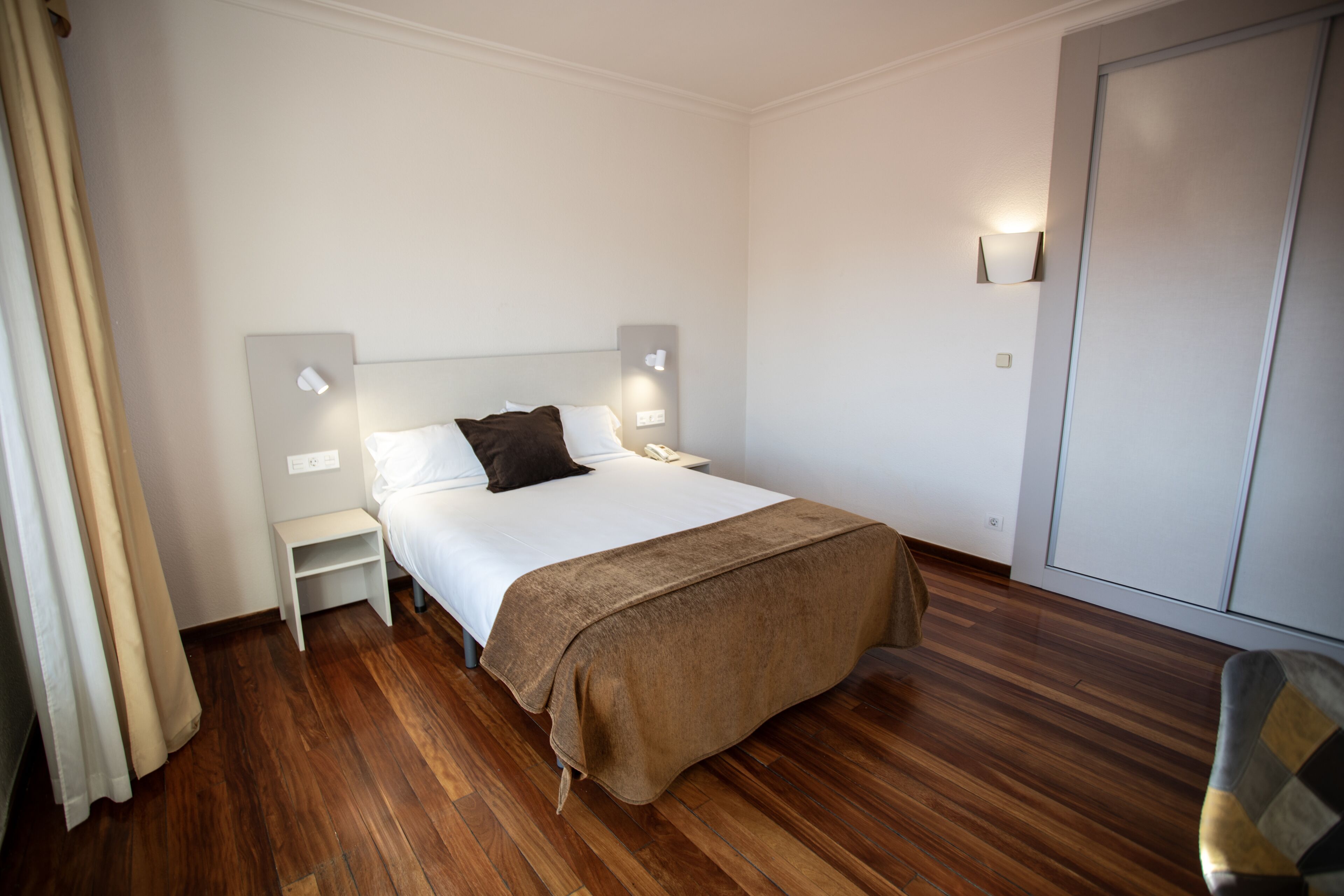 A hotel room with a single bed, a nightstand, a bedside table, a wardrobe, and a curtain.