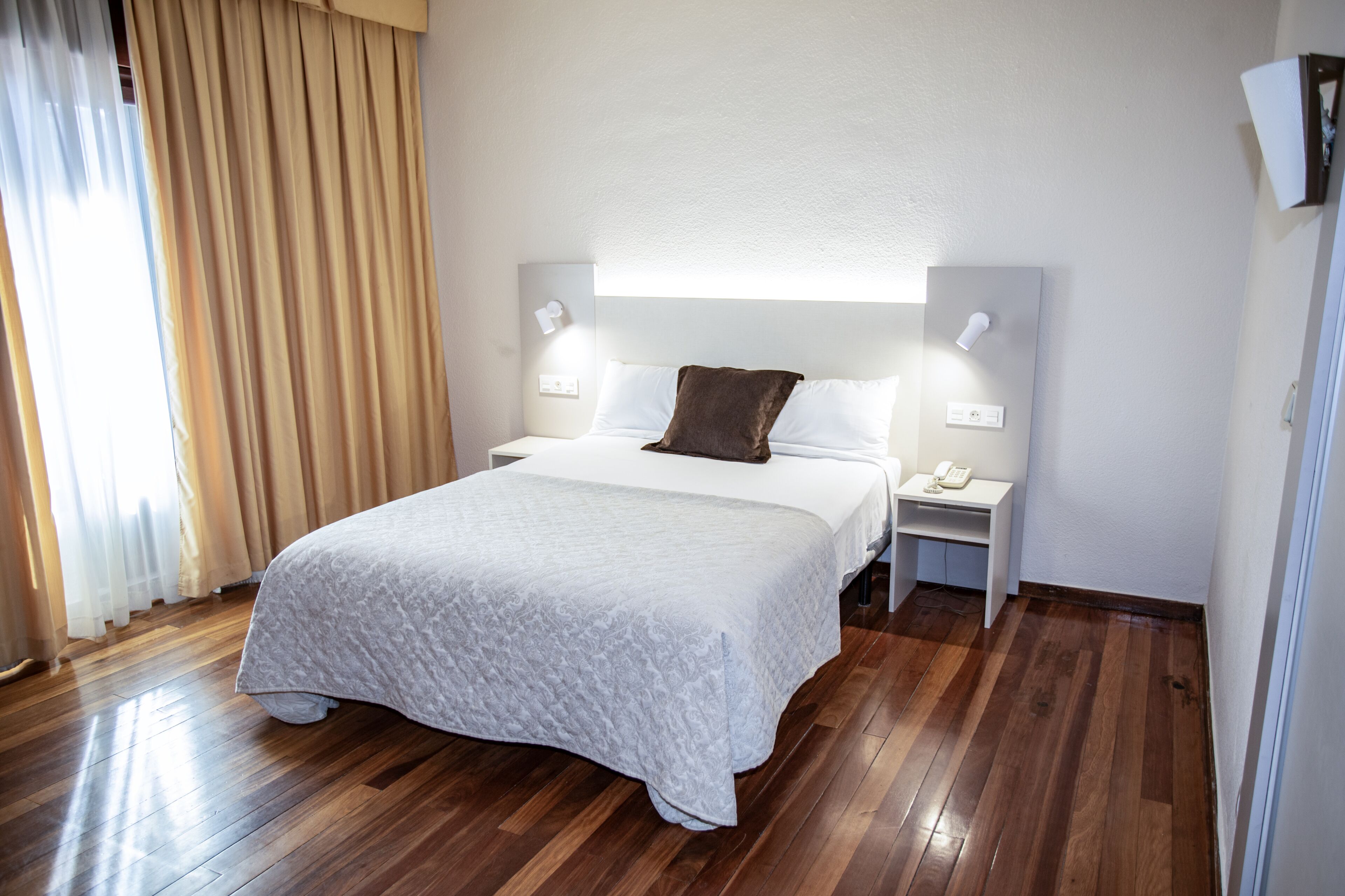 A hotel room with a bed, bedside tables, a nightstand, a lamp, a telephone, and a wall-mounted light fixture.