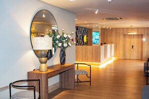 A reception area with a wooden reception desk, a mirror, a vase with flowers, and a seating area with chairs.